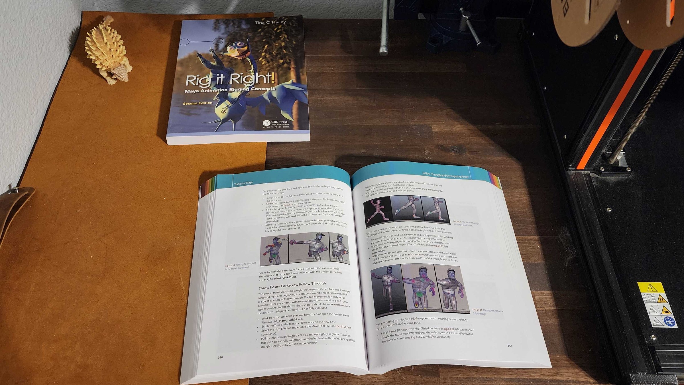 Desk with an open Maya rigging book beside a 3D printer.